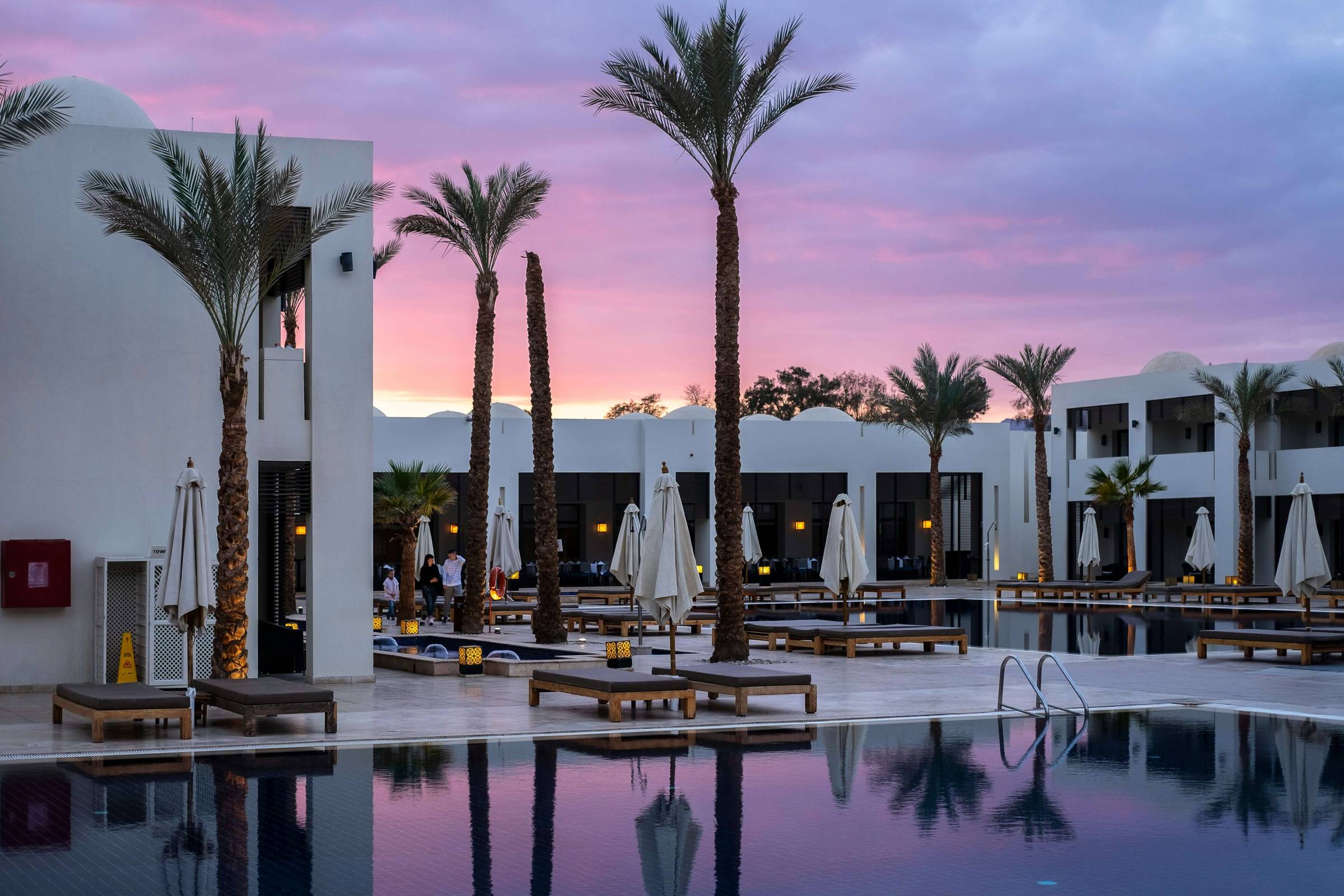 Luxury resort pool area at sunset with white modern buildings, tall palm trees, and wooden lounge chairs surrounding a calm reflective pool. The sky glows with shades of pink and purple, adding a serene ambiance to the tranquil setting.