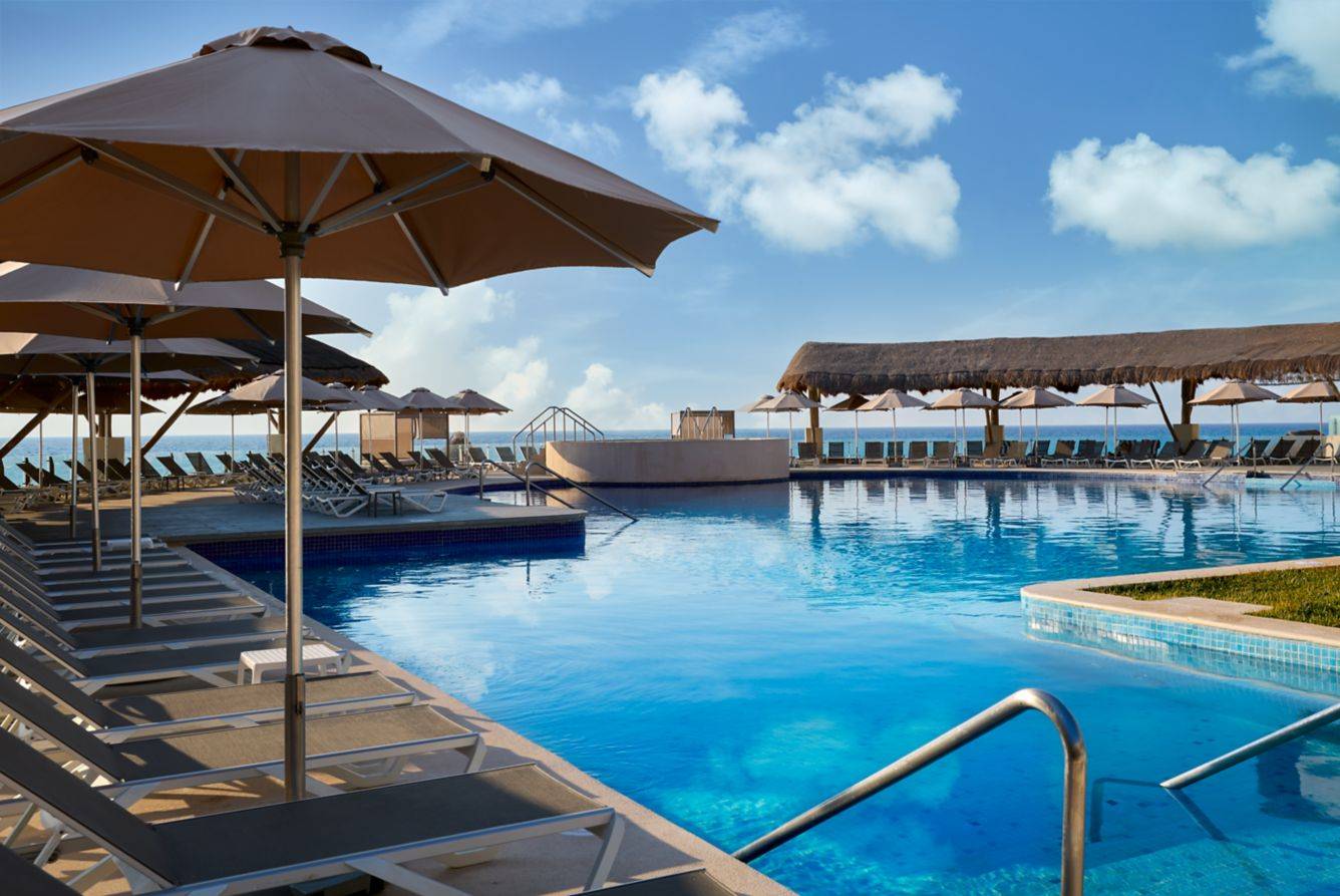 Spacious resort pool lined with lounge chairs and large umbrellas, overlooking the ocean under a clear blue sky. This luxurious beachfront setup showcases one of the ultimate travel splurges for a relaxing vacation.