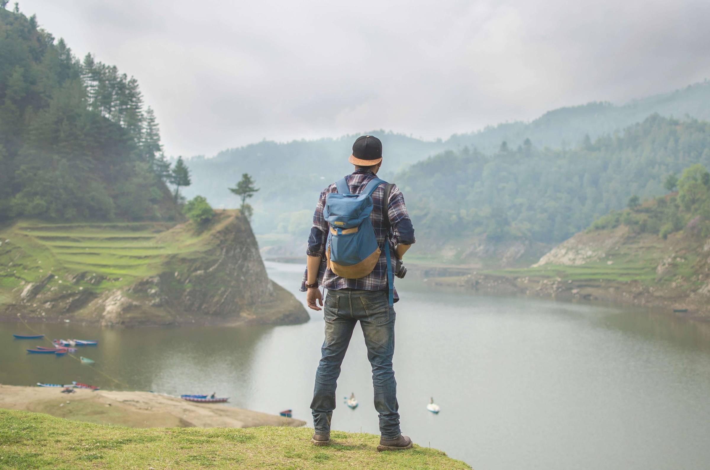 Traveler with a blue backpack and camera in hand standing on a grassy cliff, overlooking a calm river surrounded by green, forested mountains under a cloudy sky.