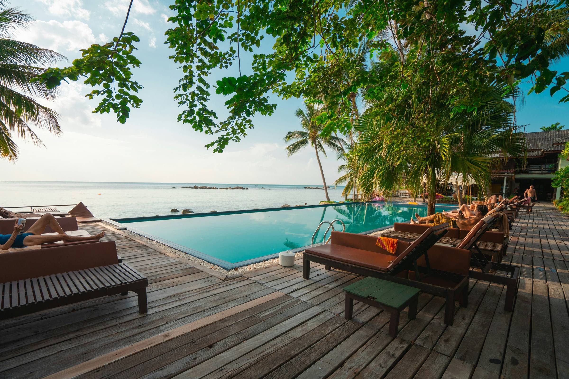 Infinity pool surrounded by wooden lounge chairs and lush palm trees overlooking a calm tropical ocean, with people sunbathing and relaxing, an ideal setting for vacation travel planning or a beach resort getaway.