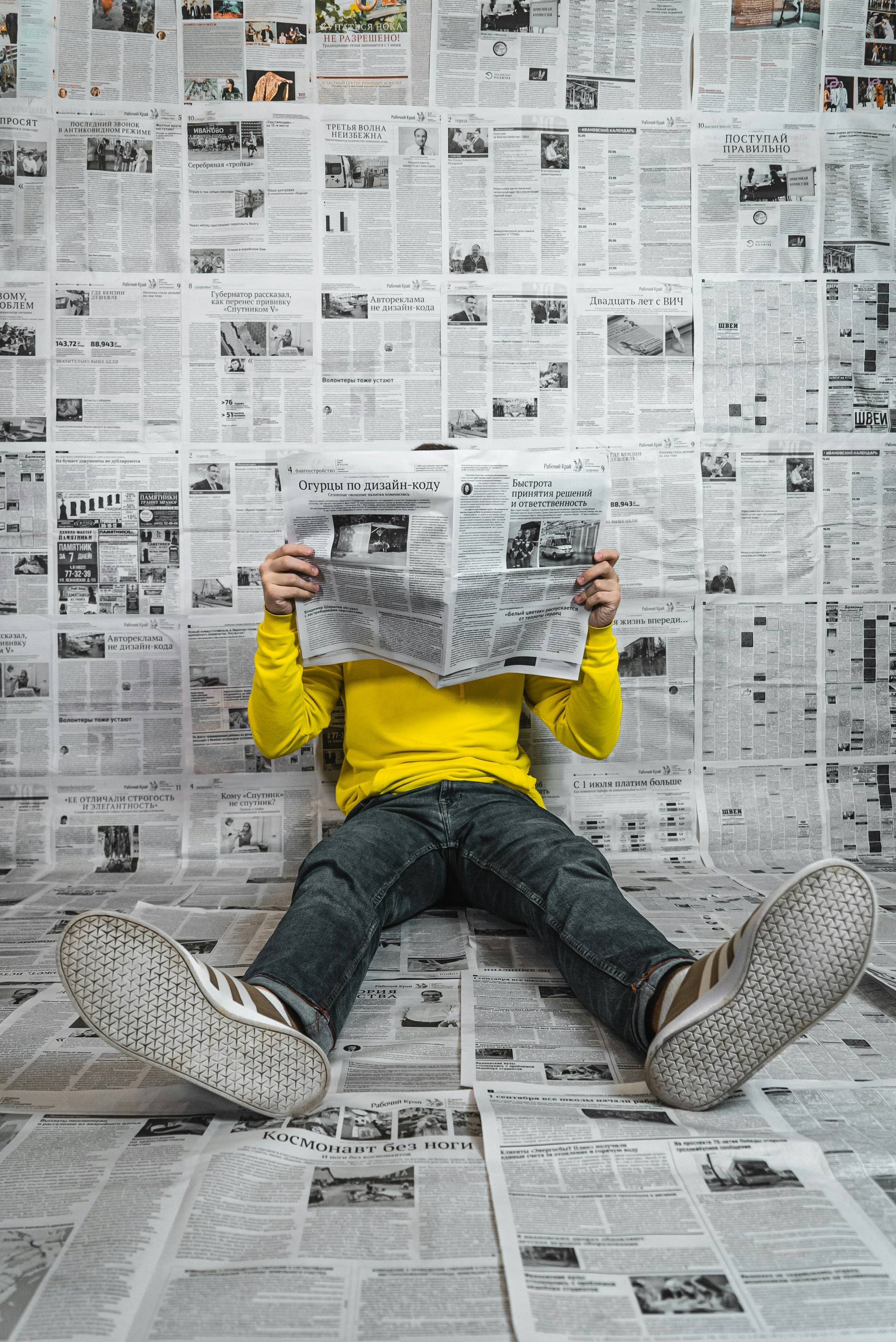 A person in a bright yellow long-sleeved shirt and jeans sits on a floor completely covered with Russian-language newspapers, reading an open newspaper that obscures their face. They're wearing white sneakers with geometric patterns on the soles. The back