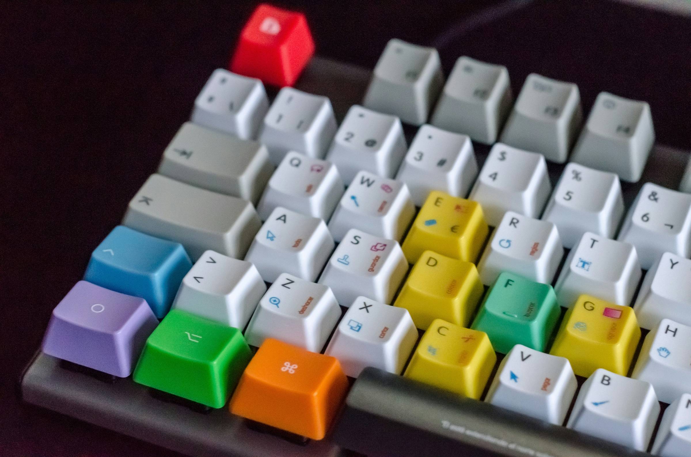 Close-up view of a mechanical keyboard with custom keycaps in various bright colors including red, yellow, green, blue, purple, and orange scattered among white keycaps, creating a colorful accent pattern against the dark keyboard base.