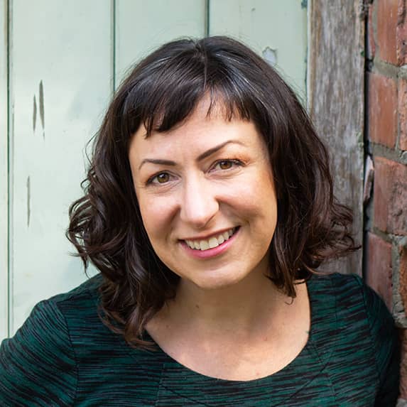 Close-up of Kim Witten, PhD. She's leaning against a brick wall, smiling, and wondering how she can help with whatever you're working on.