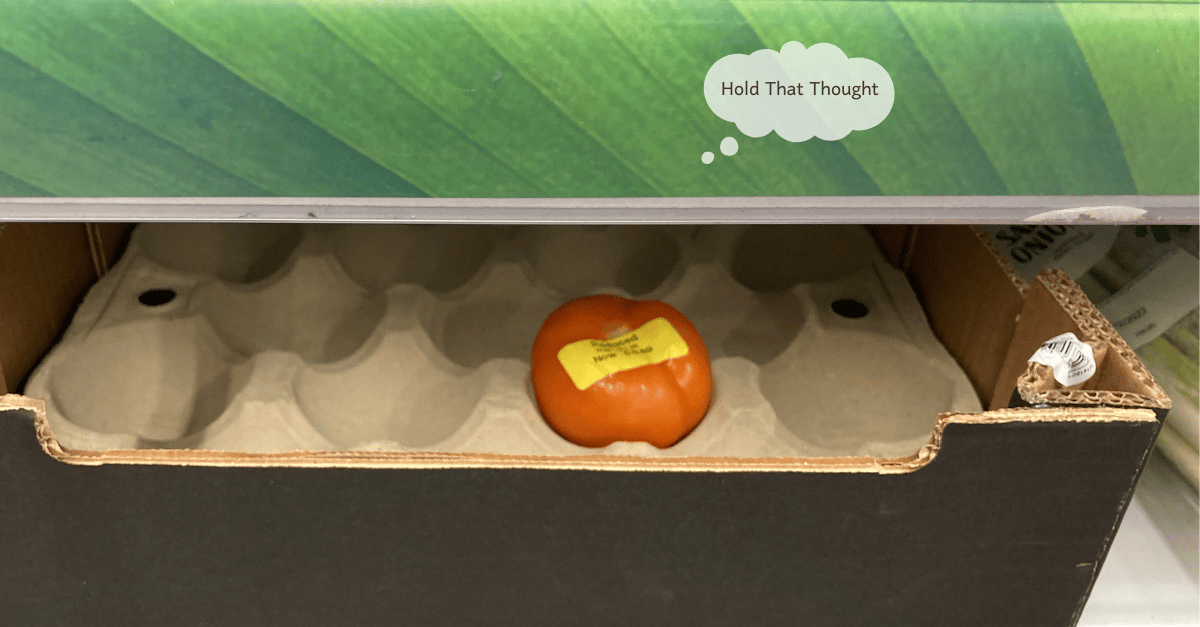 A single yellow-red tomato with a reduced sticker on it sits in a cardboard crate on a store shelf.