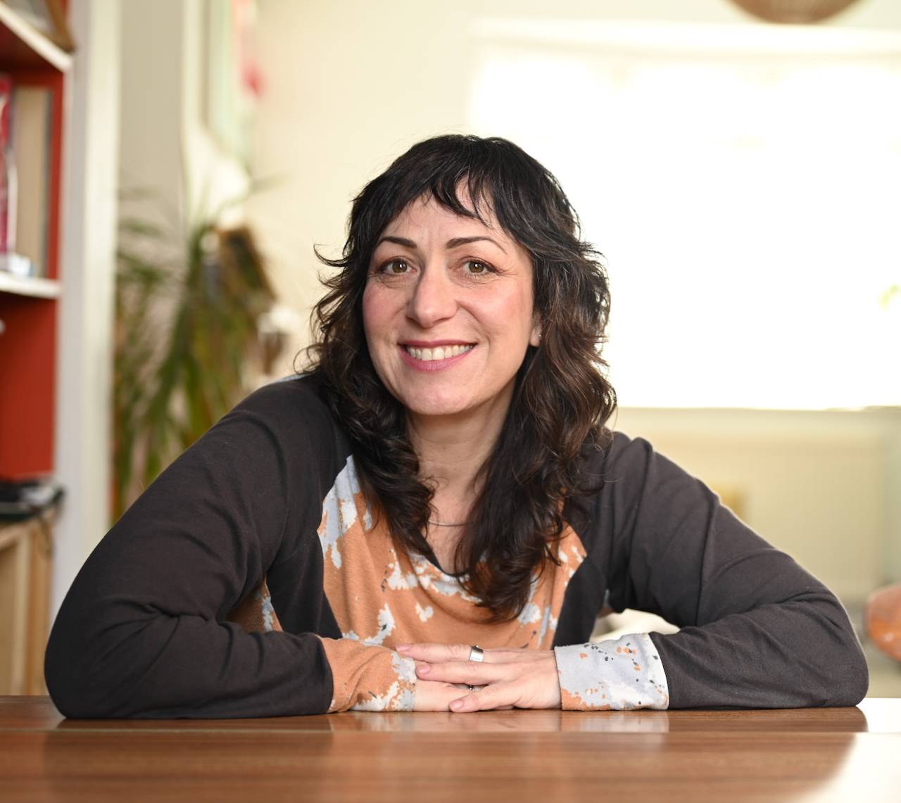 Kim has long brown hair and a colourful top she sewed herself. She's smiling at the camera with her arms folded in front of her.