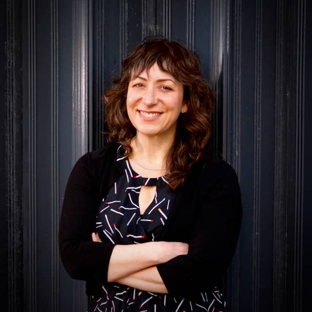 Portrait of Kim Witten, with arms folded, against a black back corrugated metal background.