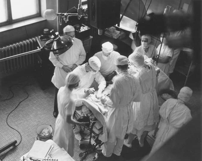 An old black and white photo of people doing surgery.  A man is standing directly behind the surgeon to direct him.  That's Vivien Thomas.