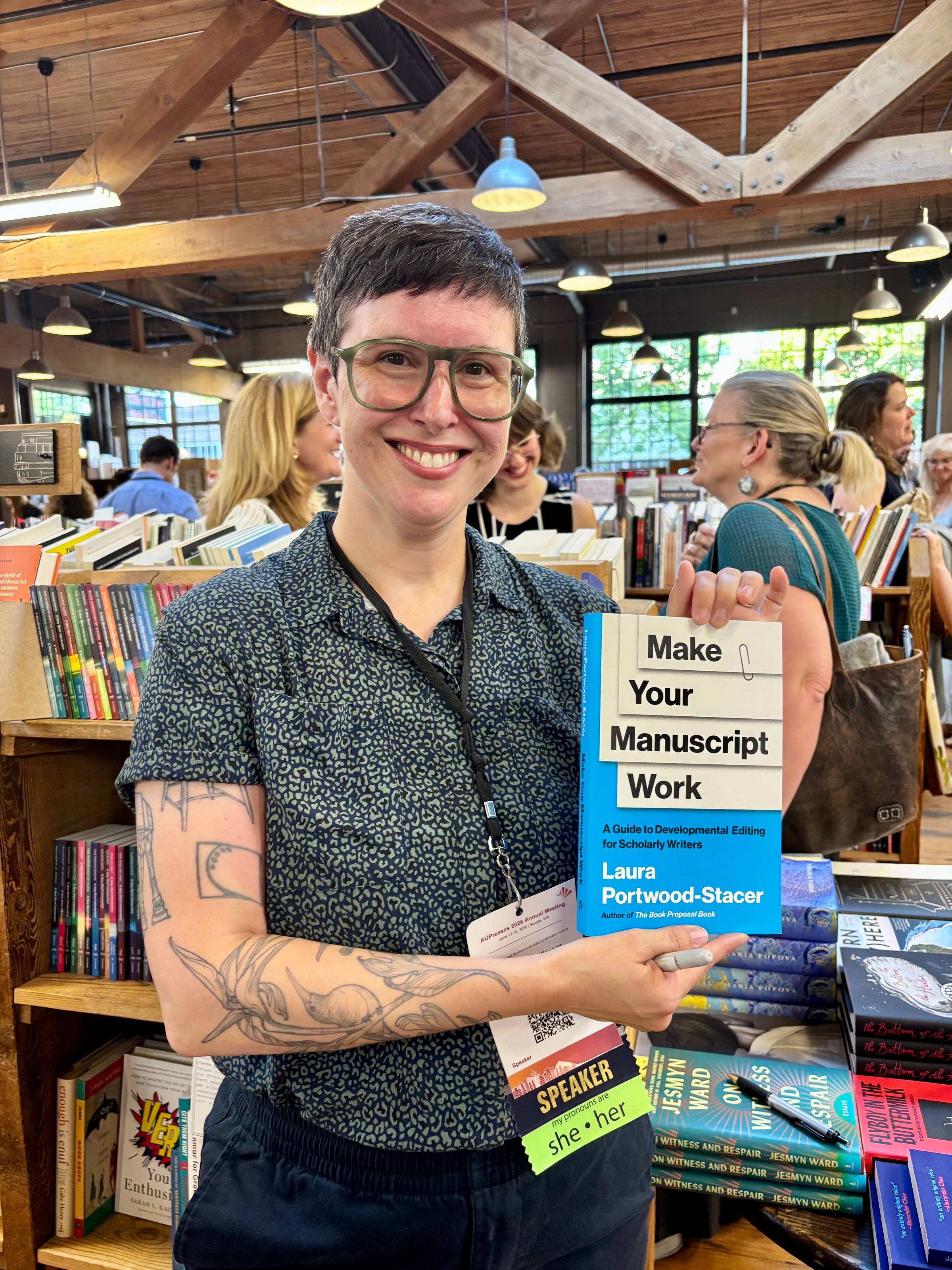 A smiling white woman with short hair, glasses, and tattoos standing in a bookstore, holding up a copy of Make Your Manuscript Work
