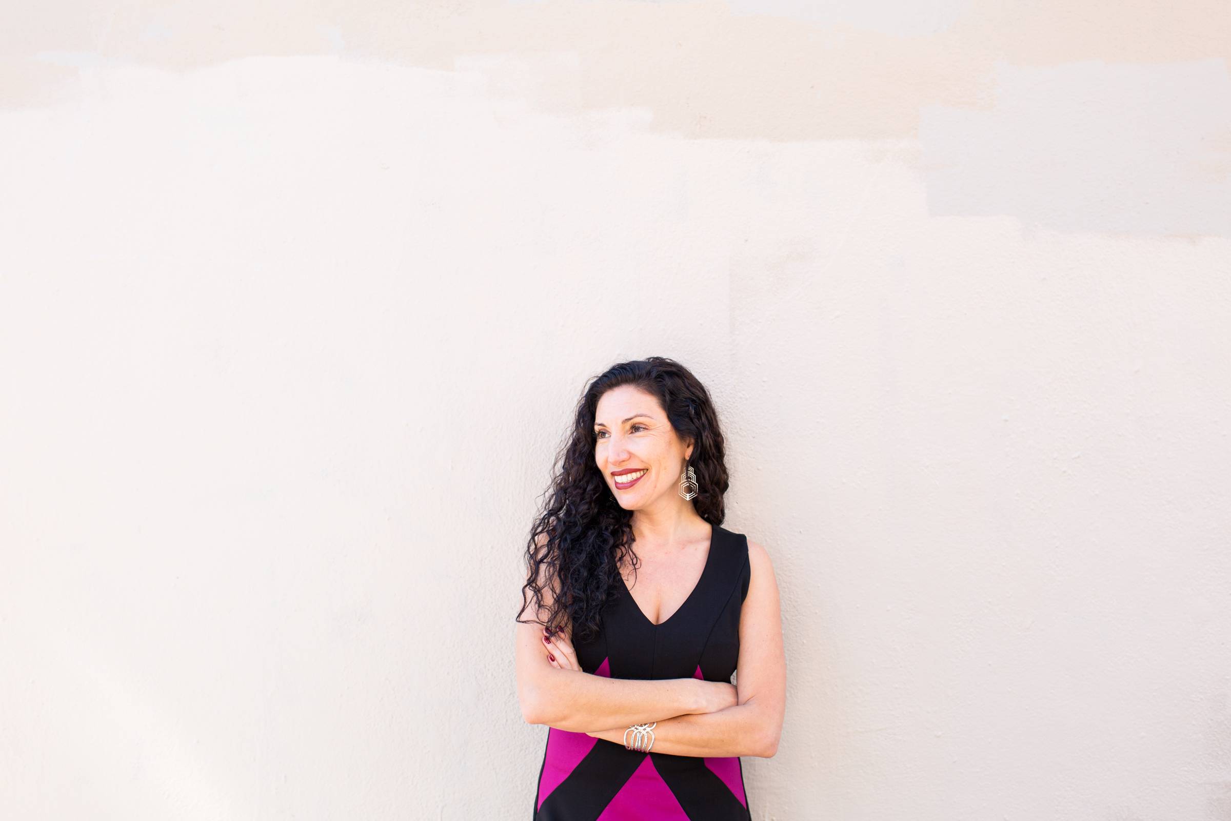 Maritza Schafer smiling confidently in a black and magenta geometric dress, arms crossed, against a warm neutral wall