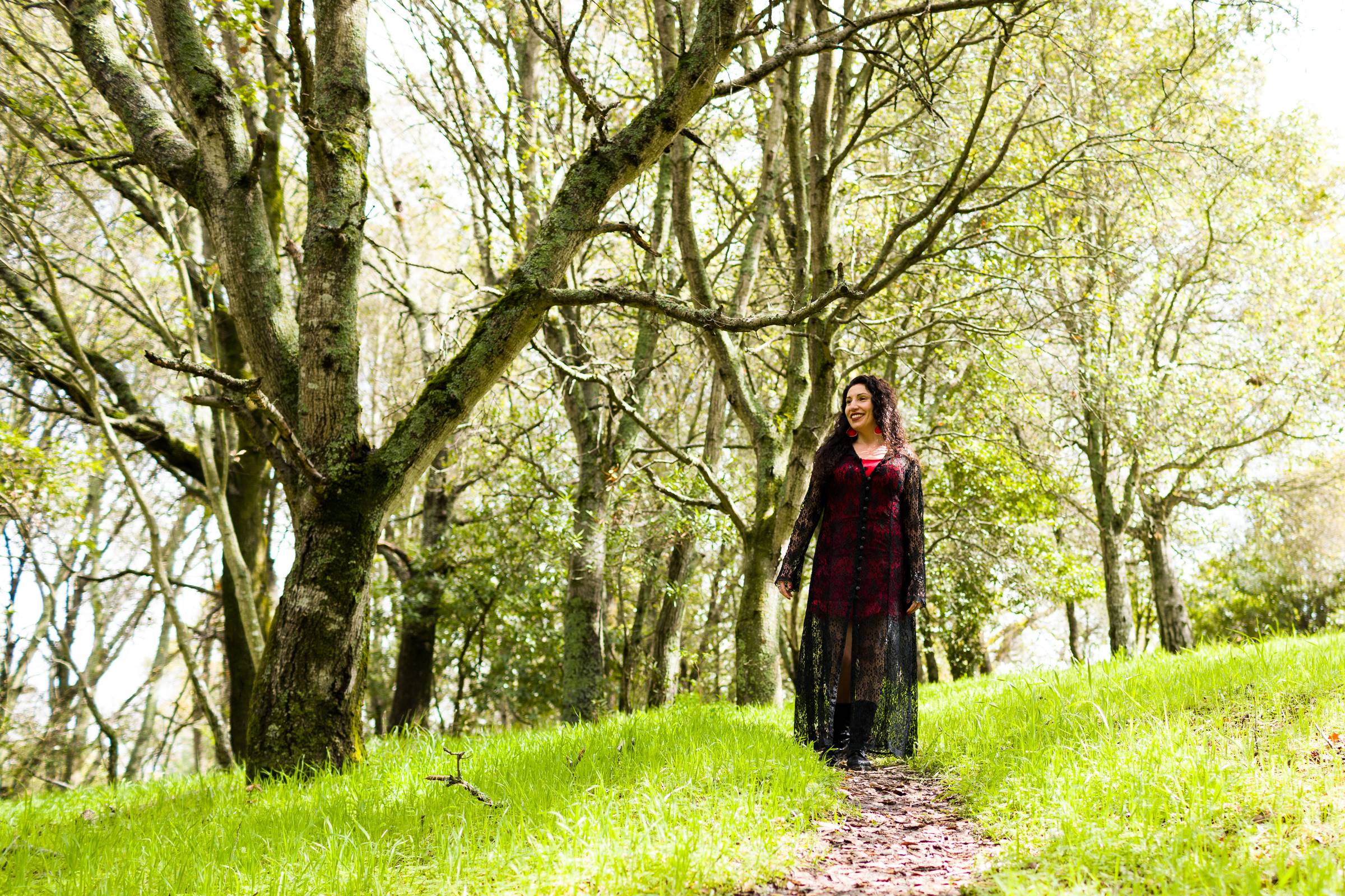 Maritza Schafer walking along a wooded forest path in a red and black lace duster, bright red earrings, smiling among sunlit trees