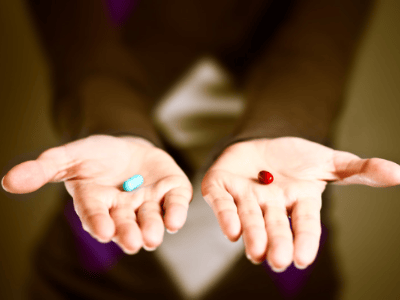 A person holding out both open palms, one with a small blue pill and the other with a small red pill, against a blurred dark background.