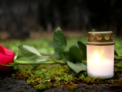 A lit white memorial candle with a gold lid sits on moss-covered ground beside a red rose and green leaves.
