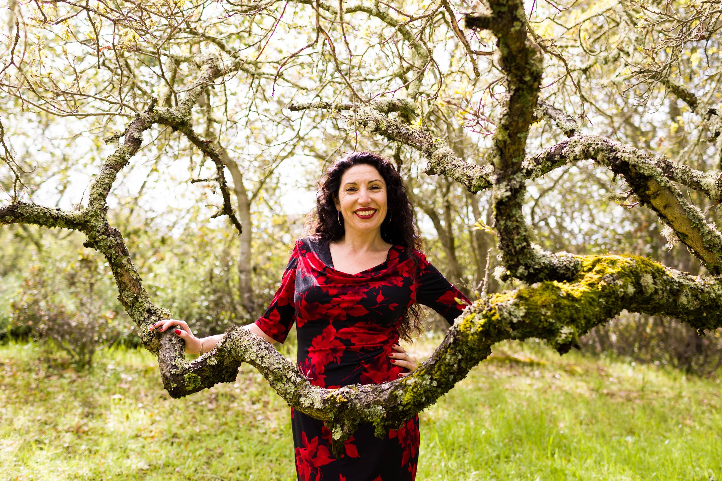 Maritza Schäfer stands between the moss-covered branches of an ancient tree in a red floral dress, smiling with quiet confidence.