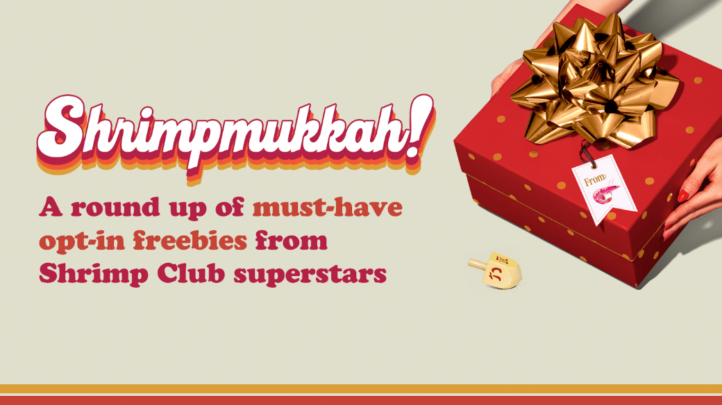 A festive promotional graphic with a light beige background. On the left, large retro-style text reads “Shrimpmukkah!” in white with a red-and-gold drop shadow. Below it, smaller red text says: “A round up of must-have opt-in freebies from Shrimp Club superstars.”  On the right side of the image, a pair of hands holds a red gift box decorated with small gold dots and topped with an oversized gold bow. Attached to the box is a gift tag that reads “From:” with a small illustration of a shrimp. Near the bottom right corner, there is a small wooden dreidel lying on its side. A thin red and gold stripe runs along the bottom edge of the graphic.