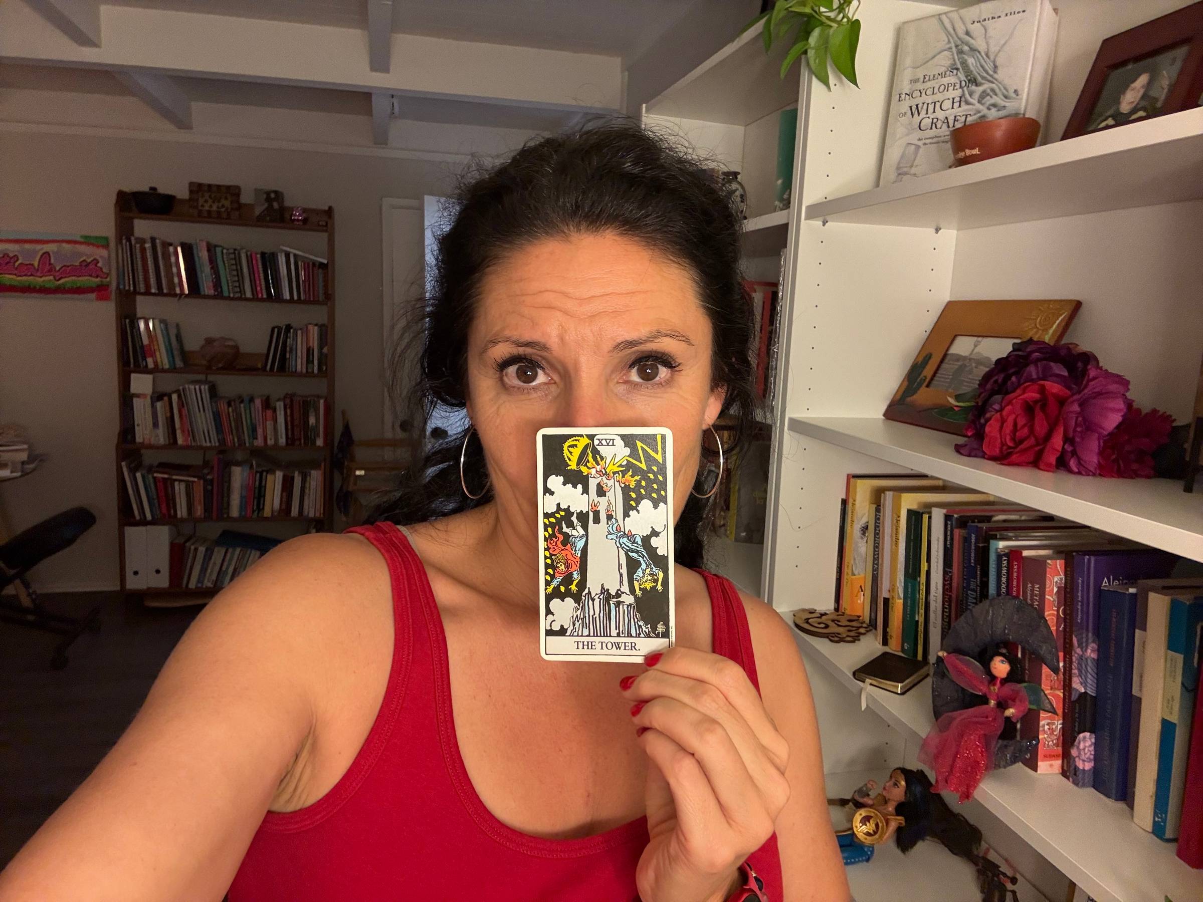 Maritza holding a Tower tarot card in front of her face, standing in front of a bookshelf filled with books and spiritual objects.