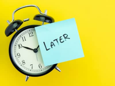 Black alarm clock with a light blue sticky note reading "Later" stuck over its face, set against a bright yellow background.