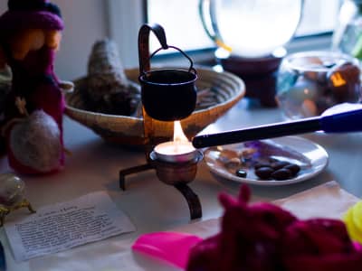Small cauldron ritual kit being lit with a torch lighter, surrounded by dried herbs, a felt gnome figure, tumbled stones, and pink flower petals on a witchcraft altar setup