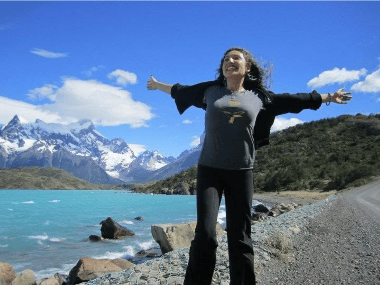 Maritza Schafer with arms raised in celebration on a shore, with turquoise lake and snow-capped mountains in the background.