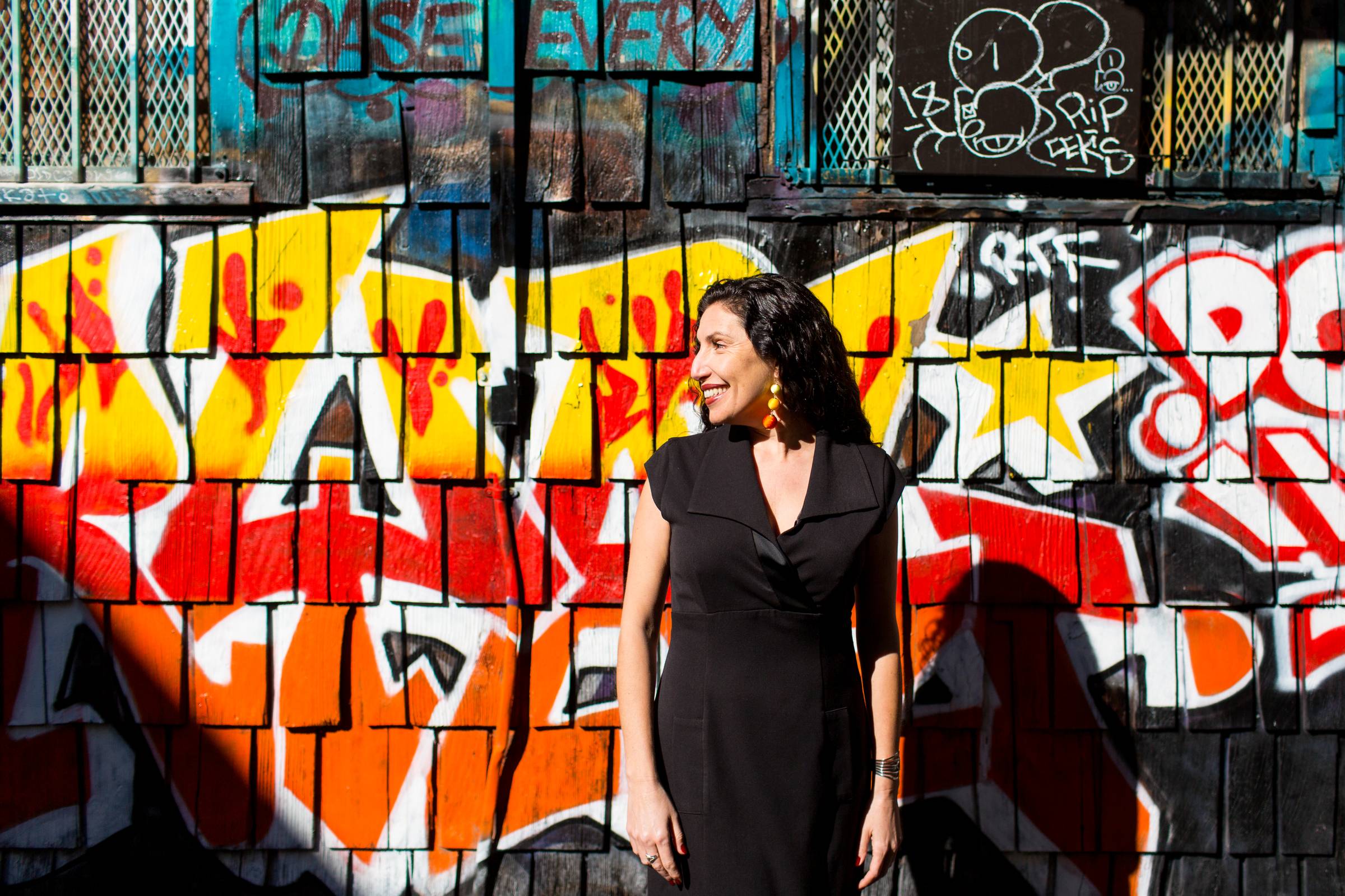 Maritza Schäfer smiling in a black dress against a colorful red, yellow, and orange graffiti wall.