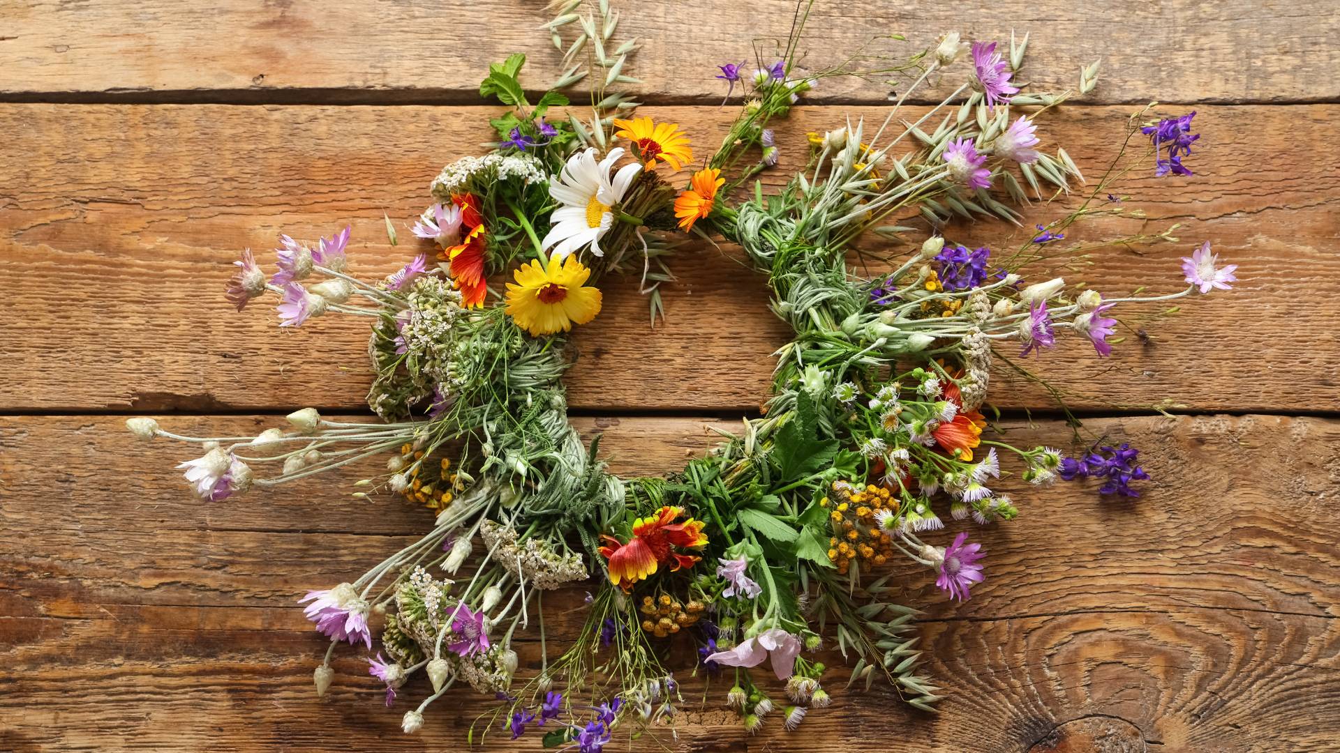 A wildflower crown laid flat on a weathered wooden surface, woven from fresh herbs and greenery and dotted with blooms in yellow, orange, red, white, and purple, including calendula, daisies, and small violet flowers.