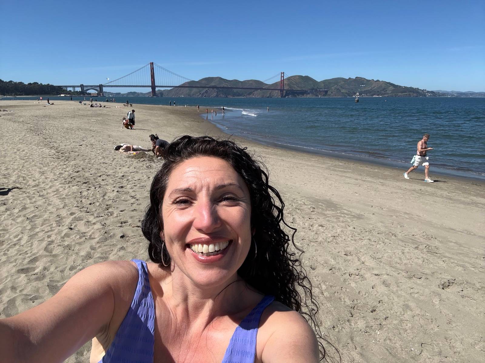Maritza smiling in a beach selfie with the Golden Gate Bridge behind her, people walking along the shoreline under a clear blue sky.