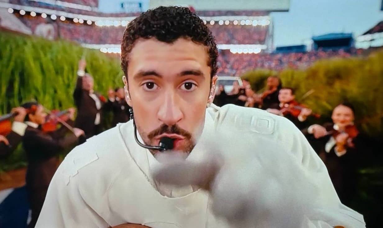 Television screen showing a close-up of Bad Bunny performing during the Super Bowl,  wearing a white outfit and a headset microphone, leaning toward the camera with an intense, focused expression. An orchestra plays behind him in a stadium setting, with b