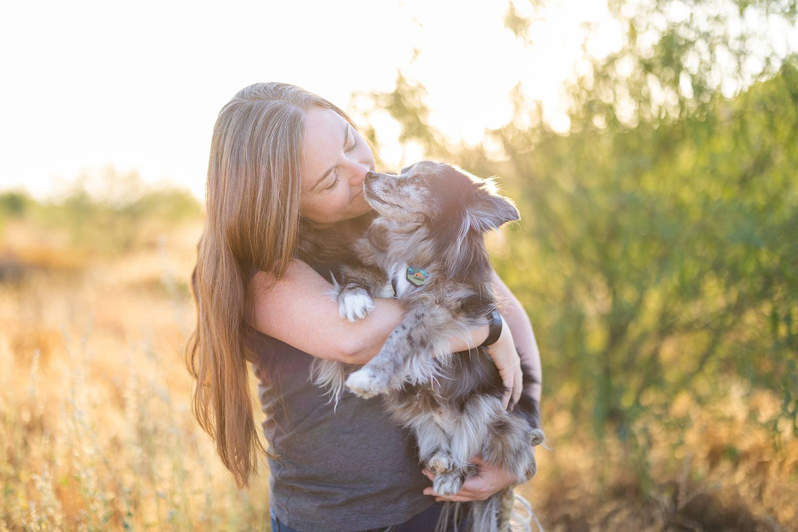 Sara, a white woman with long brown hair, stands in a desert setting at sunset holding her heart dog Finnick, a small brown and black fluffy dog, as they gaze at each other with deep love