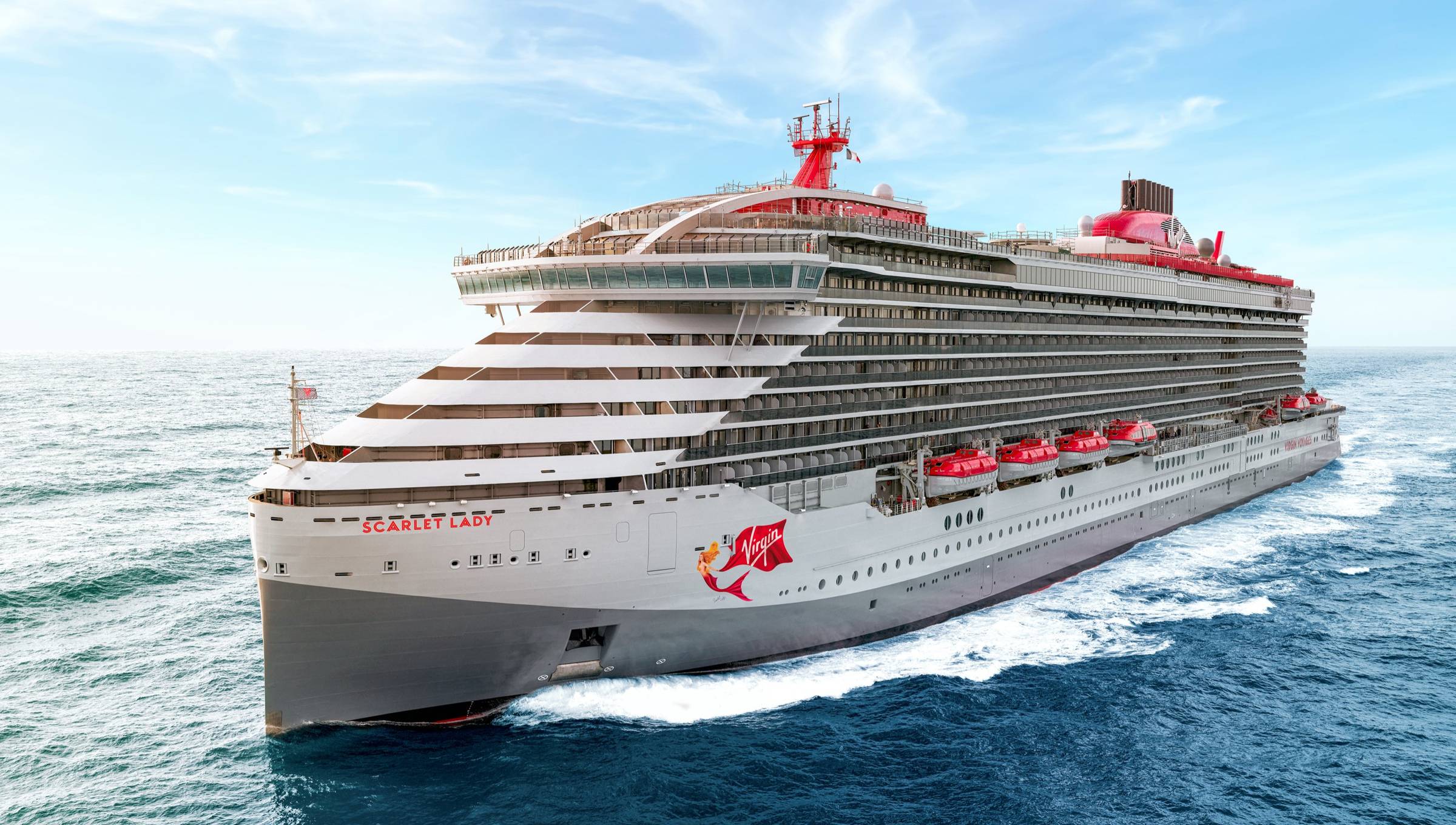 Virgin Voyages Scarlet Lady, shown from a front-side angle, with a sleek gray hull, red accents, and the Virgin logo painted on the side.
