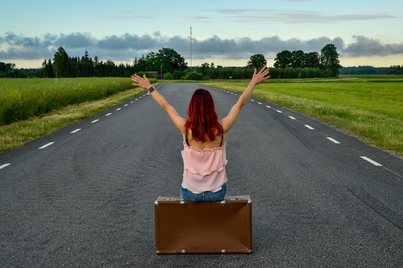 Woman with raised arms sitting on suitcase on open road.