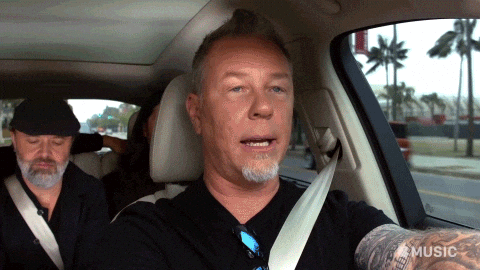 James Hetfield show a rock on (horns) hand sign while driving in a car. LArs Ulrich can be seen on the backseat.