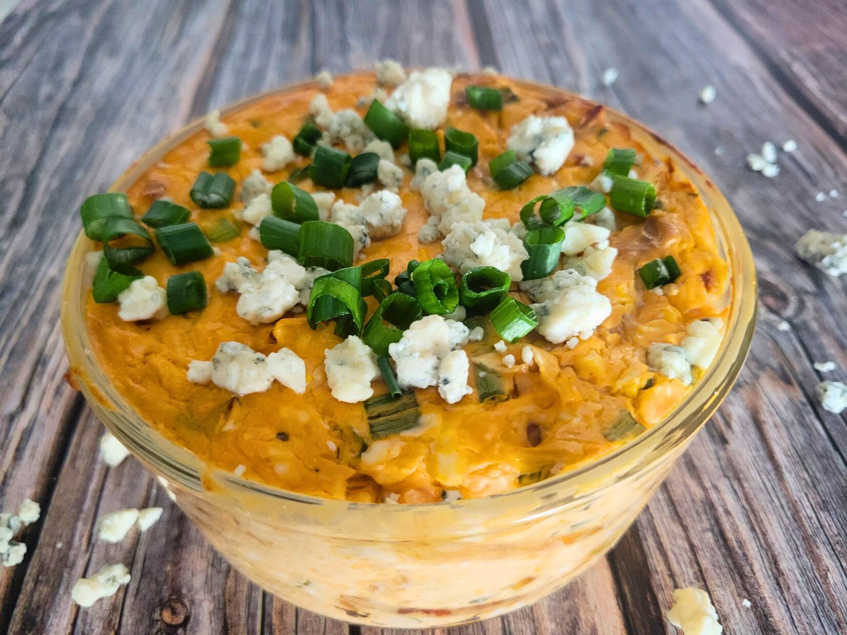 jackfruit buffalo dip garnished with green onions and blue cheese crumbles in a clear bowl on a wooden surface.