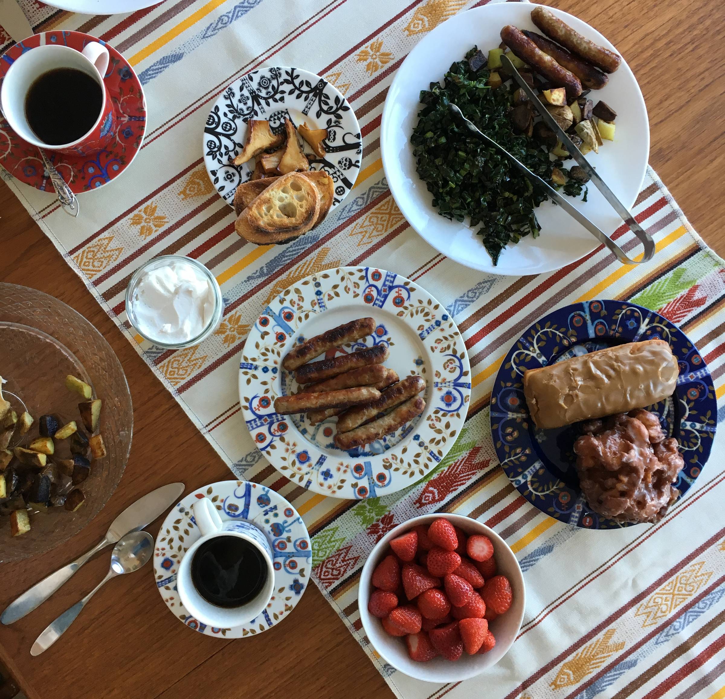 A wood table with a colorful table runner, two cups of coffee, and a spread of sausages, potatoes, kale, donuts, and strawberries.