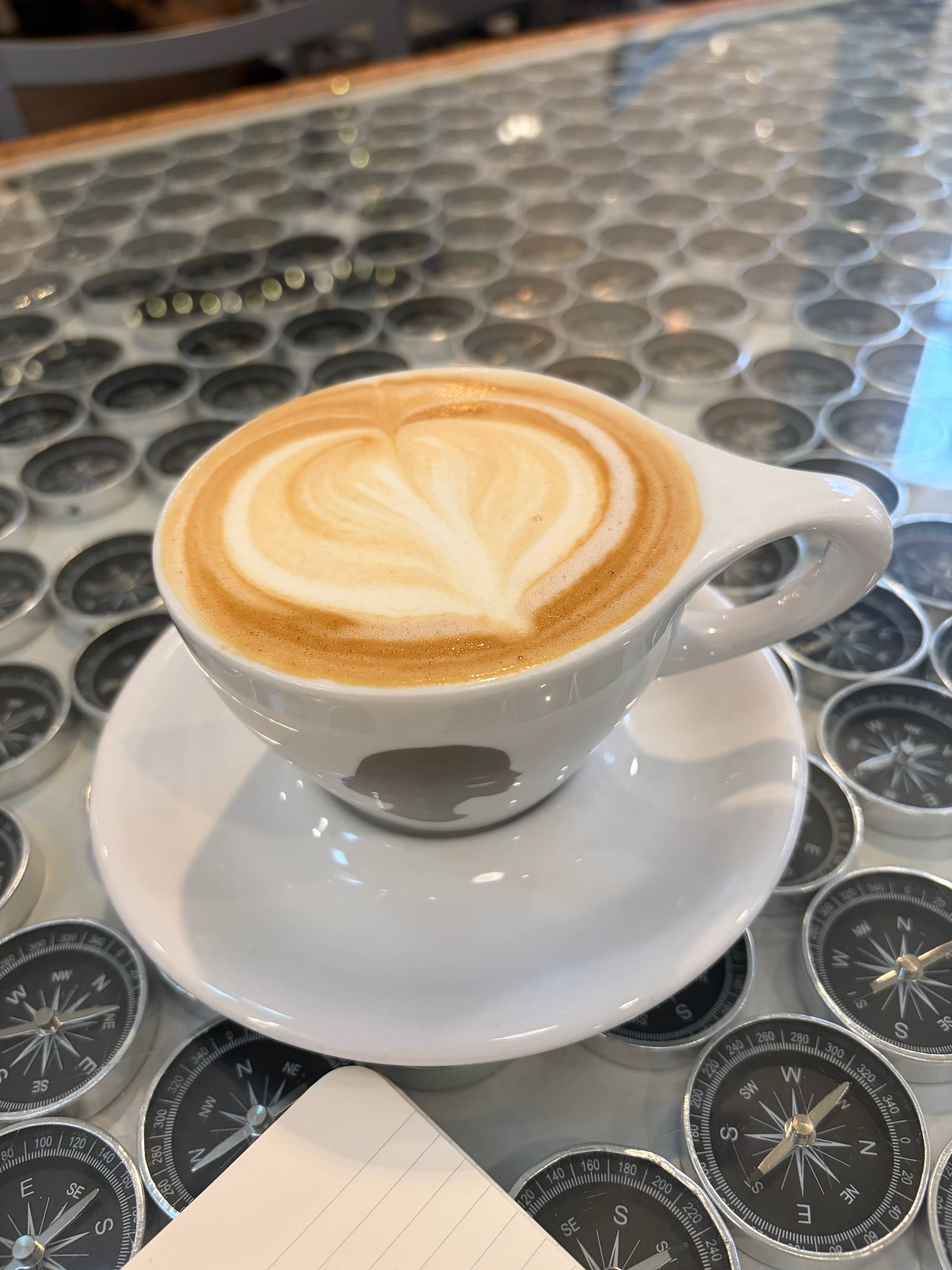 A shadowbox table filled with hundreds of tiny compasses beneath glass, a cappuccino and the corner of writing paper visible.