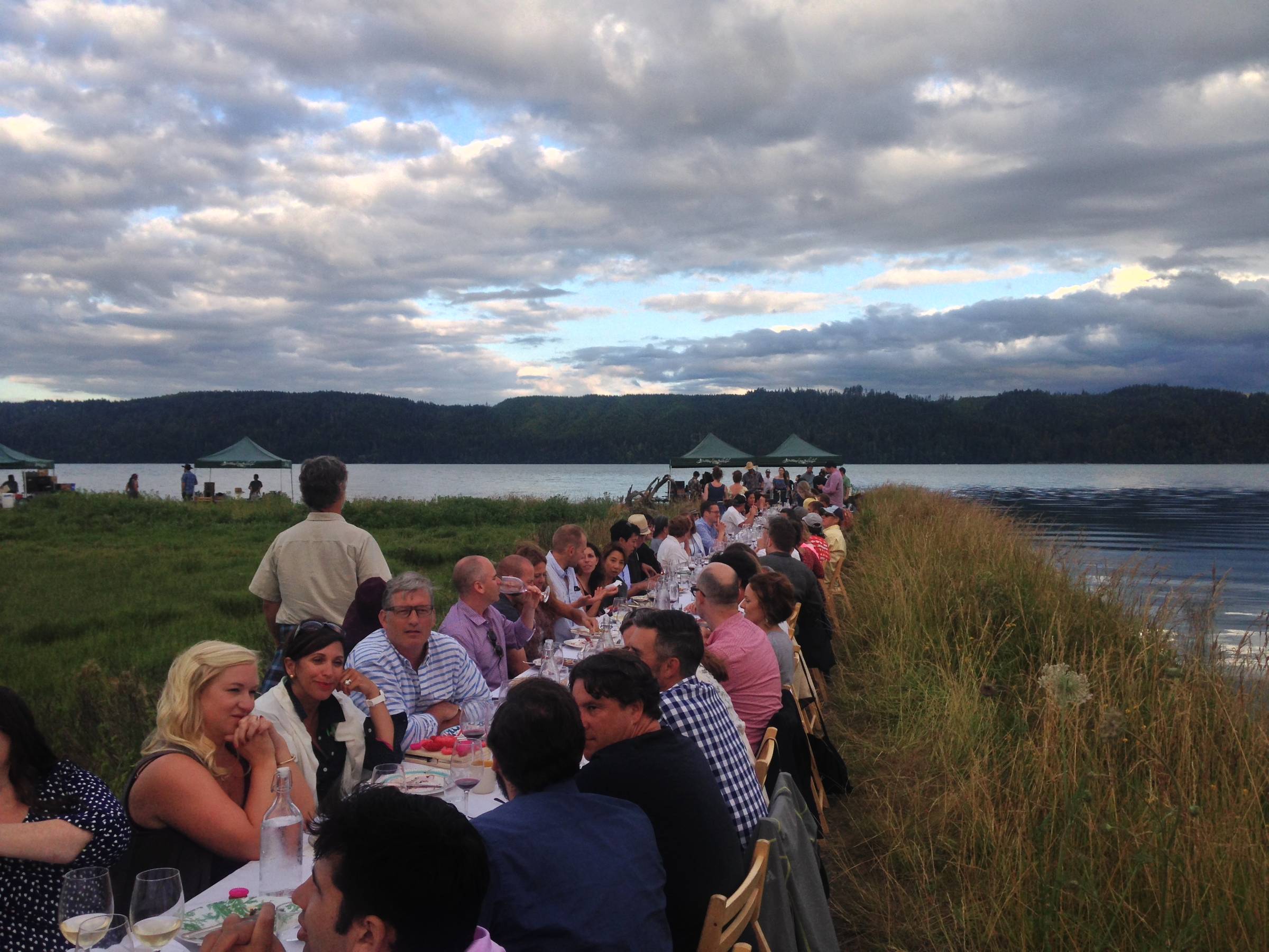Diners seated at a long communal table beside calm open water, the table stretching away from the viewer.