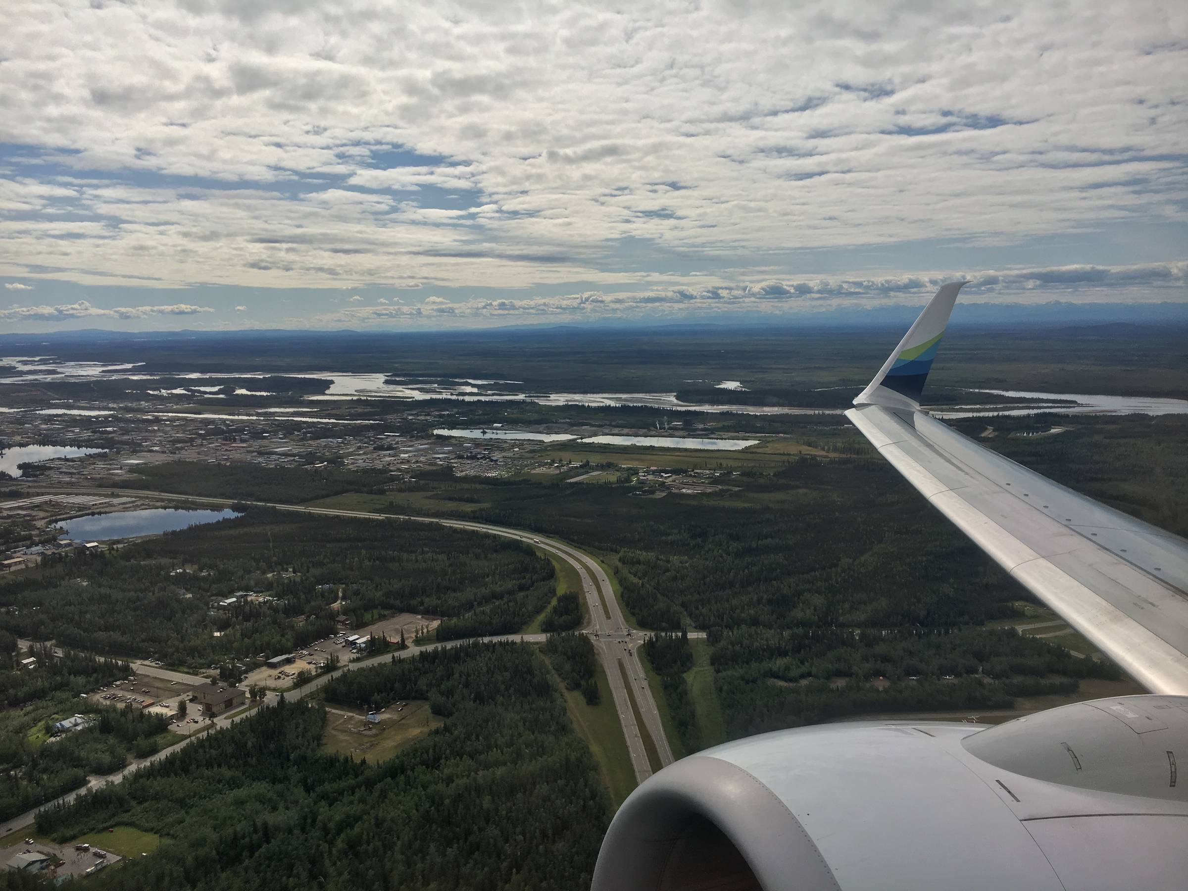 Airplane wing and engine over Alaska terrain with lakes, roads, and summer clouds