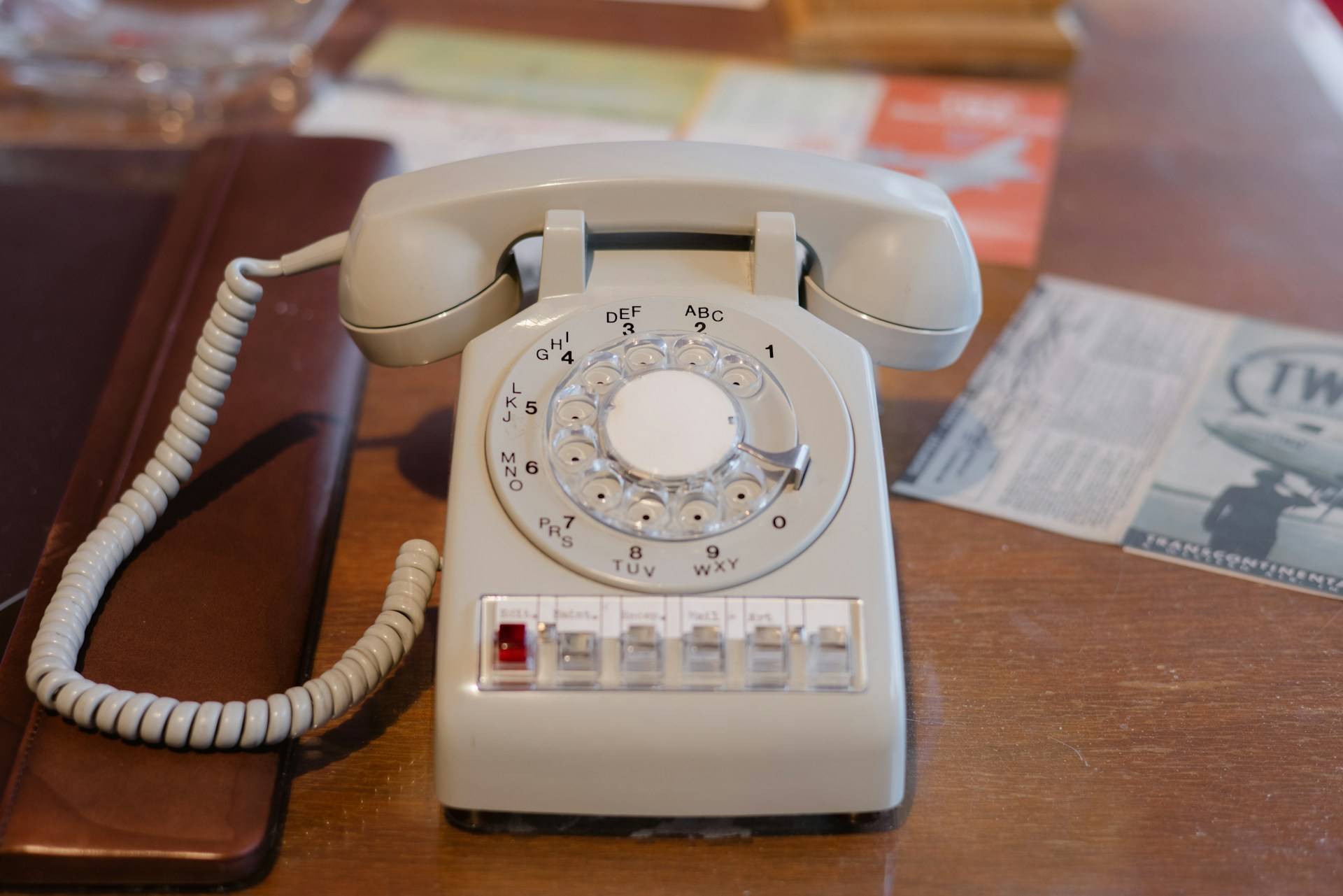 Vintage tan rotary phone on wooden desk with scattered papers and leather folio pad.