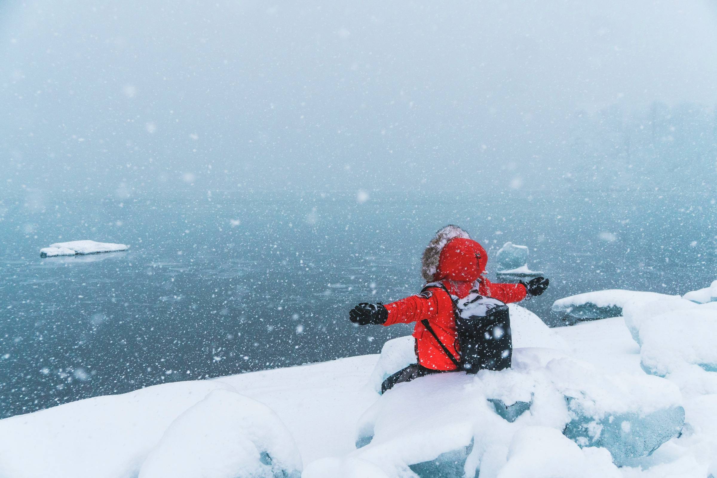 Person in red jacket sits on snowy bank, arms outstretched, facing icy water and falling snow