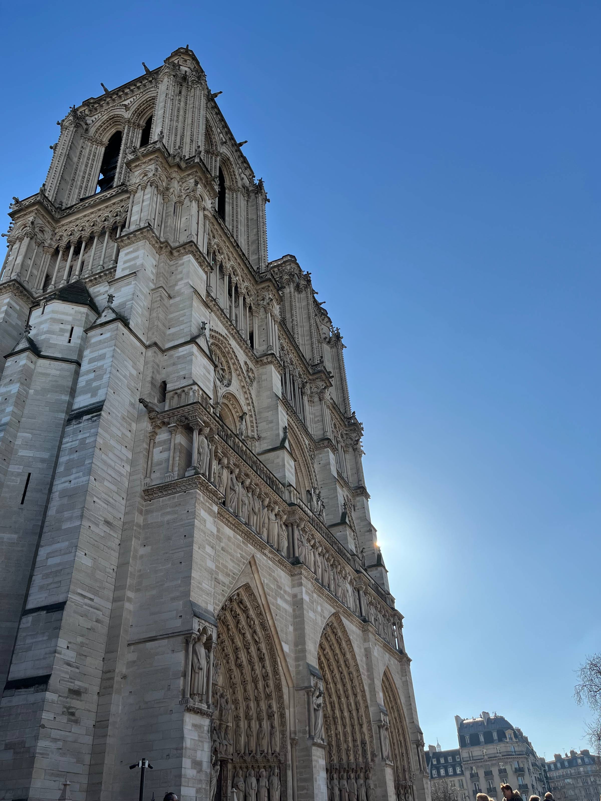 Notre Dame's stone facade backlit by sun, a bright point of light breaking through against a blue sky.