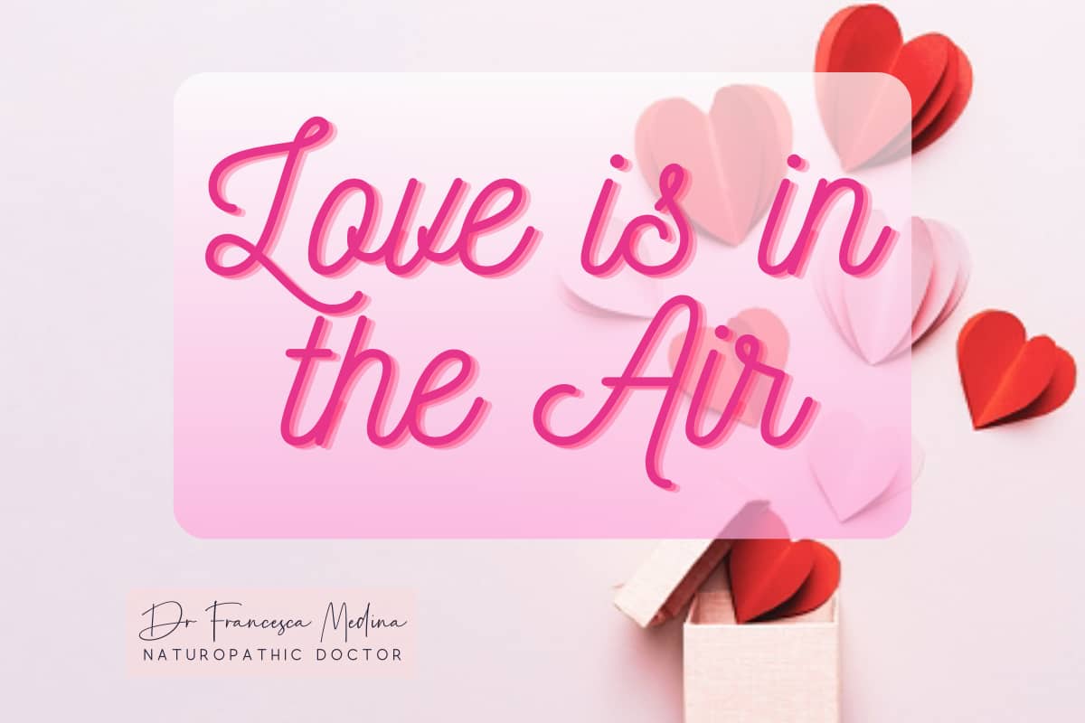 The image features the phrase "Love is in the Air" written in pink, cursive letters against a light pink background. Behind the text, there are various red and pink paper hearts scattered decoratively. In the bottom left corner, there is a signature-like 