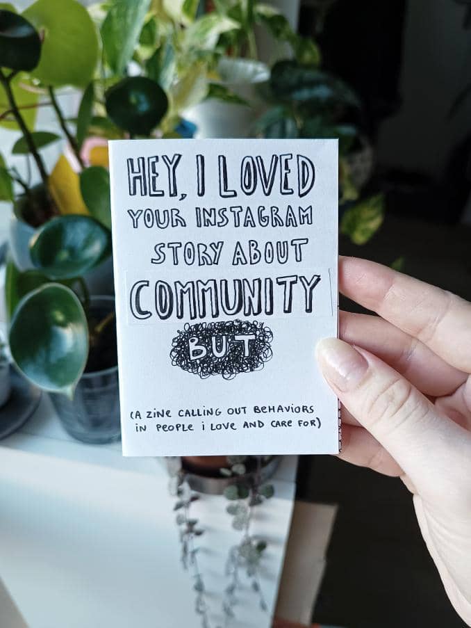 A hand holding up the paper zine, which reads Hey, I loved your story about community but (a zine calling out behaviors in people I love and care for)