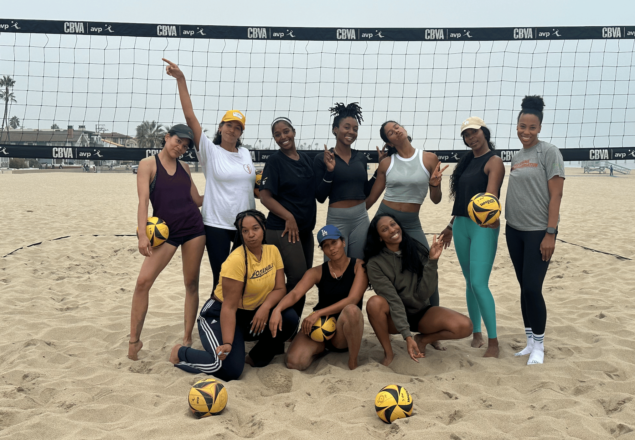 Black women's beach volleyball community gathering on Manhattan Beach court, with players holding yellow volleyballs and posing together after their first meetup