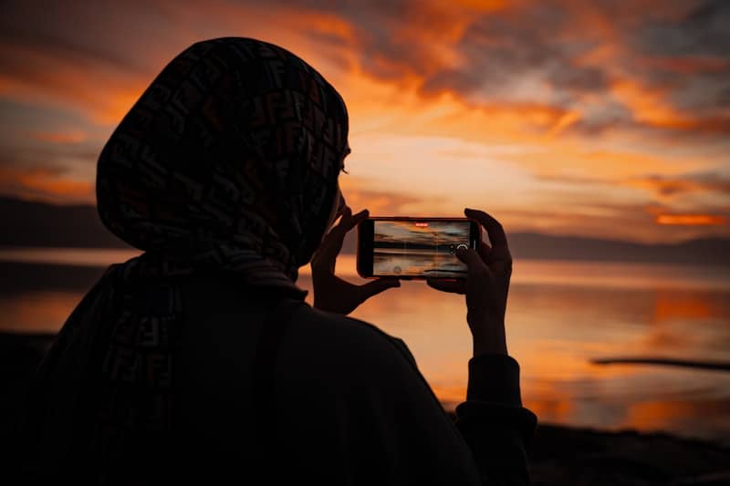 Woman uses mobile phone to photograph vibrant sunset over calm water