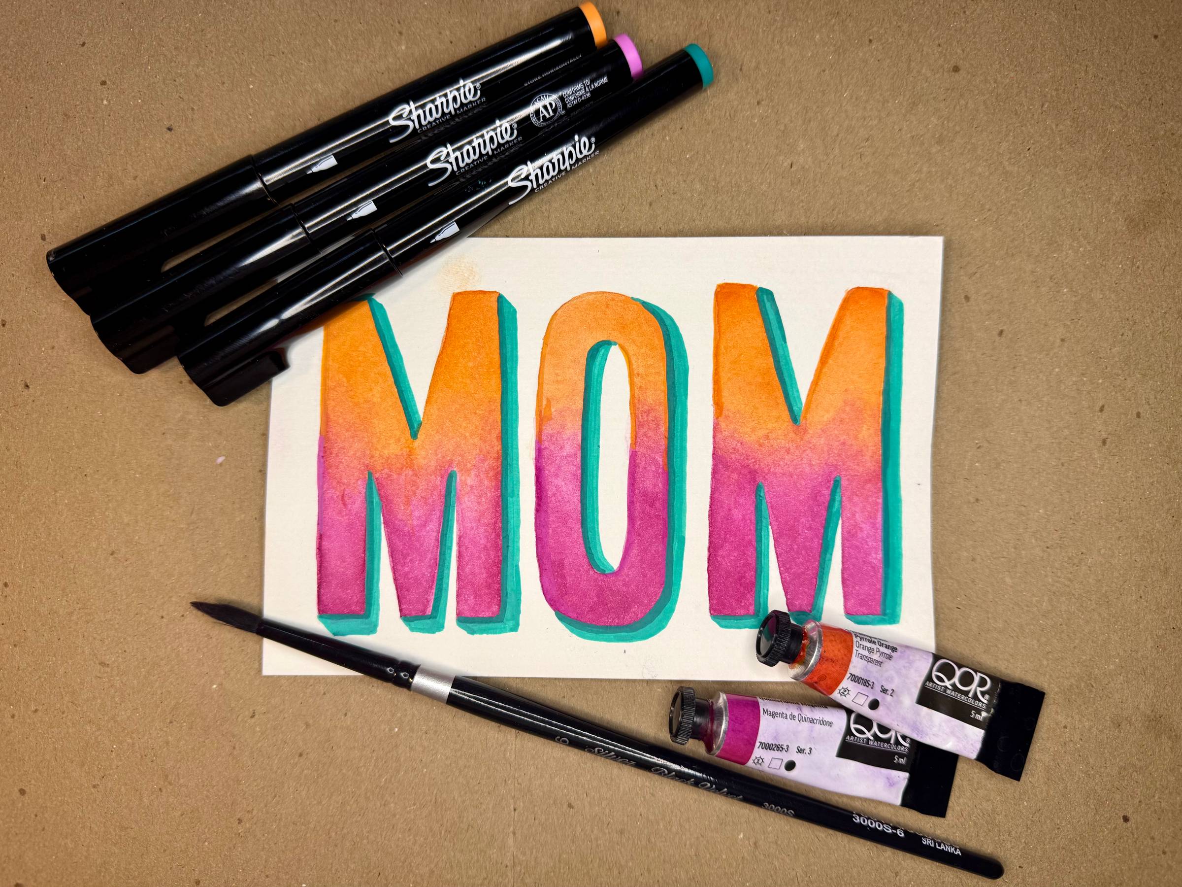 MOM in block letters painted in with watercolors in pink and orange with a shadow in turquoise pen.