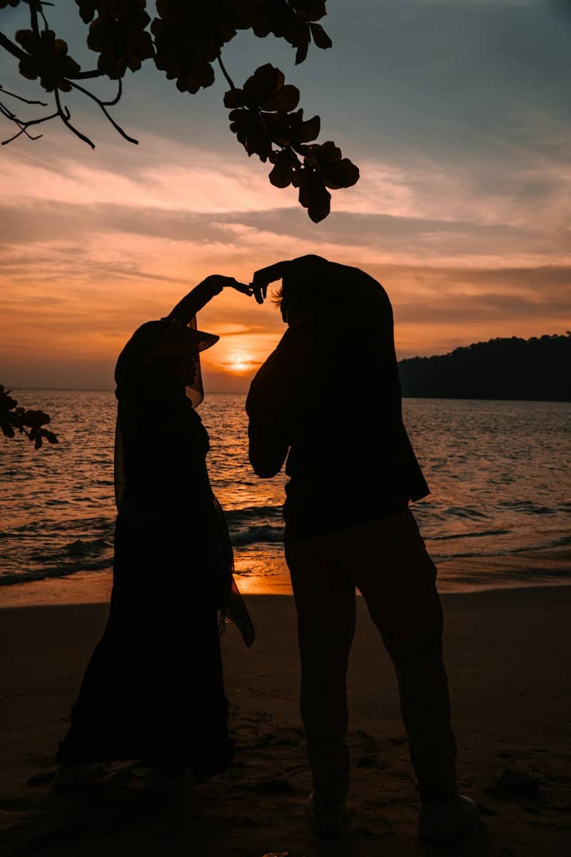 Two people in silhouette forming a heart shape with their arms on a beach