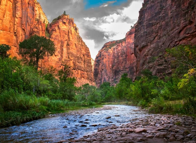 brown rocky mountain beside green trees under white clouds and blue sky during daytime