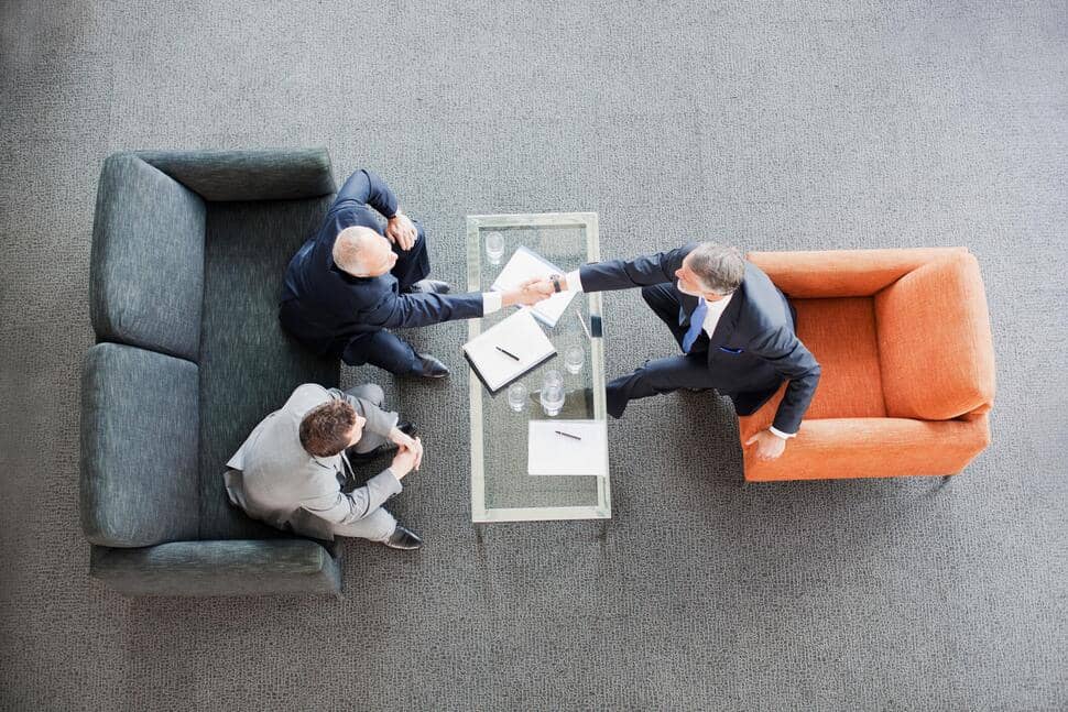 Overhead shot of two men sitting on one side of a coffee table and shaking the hand of one man sitting on other side of table, like in an interview