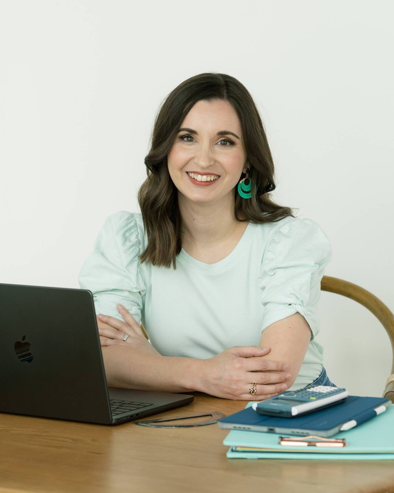 A person with shoulder-length dark hair, wearing a light green blouse, is sitting at a wooden desk. There is a black laptop in front of them. On the desk, there are a stack of notebooks, a pen, and a calculator. The person is smiling and looking at the camera. The background is plain white.