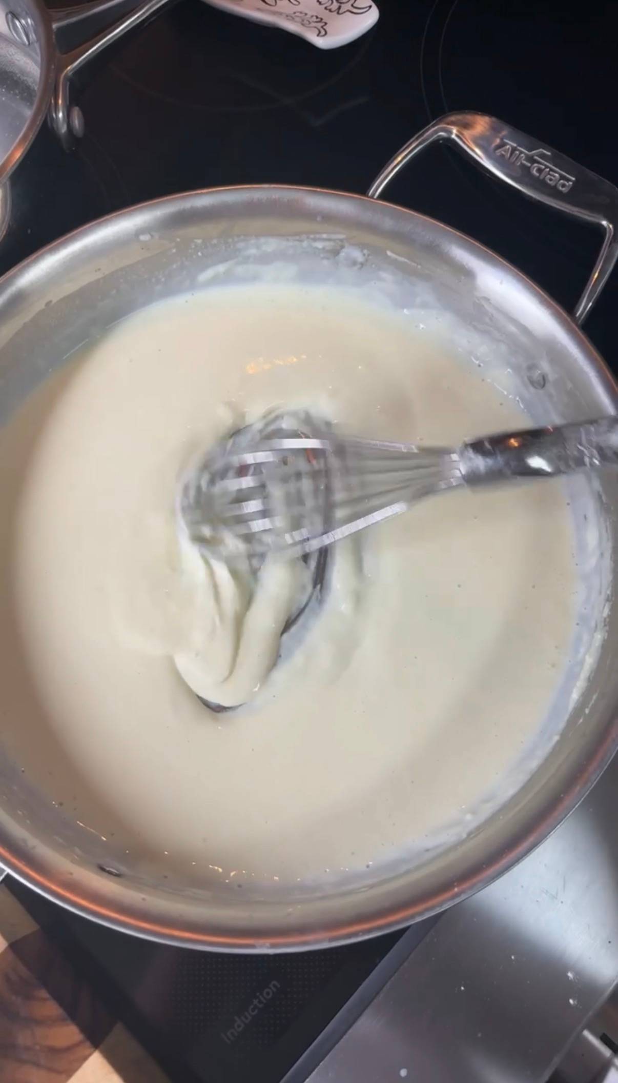 Overhead view of smooth, finished béchamel sauce in a stainless All-Clad saucepan, whisk resting in the creamy white sauce