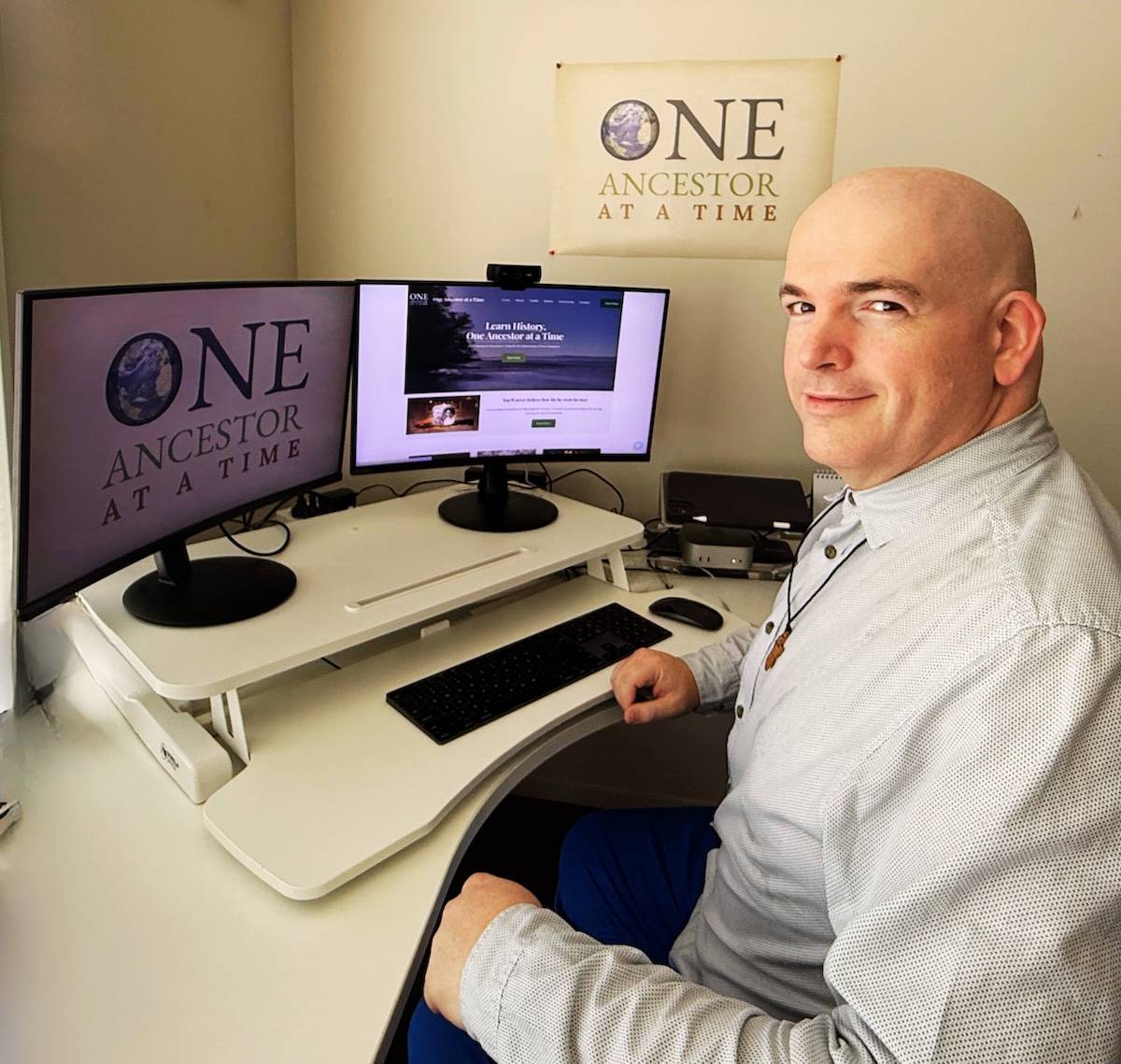 A person sitting at a desk with dual computer monitors, both displaying the text "ONE ANCESTOR AT A TIME." The individual is smiling at the camera, wearing a light-colored shirt. On the wall behind them is a poster with the same text. The desk has a keybo