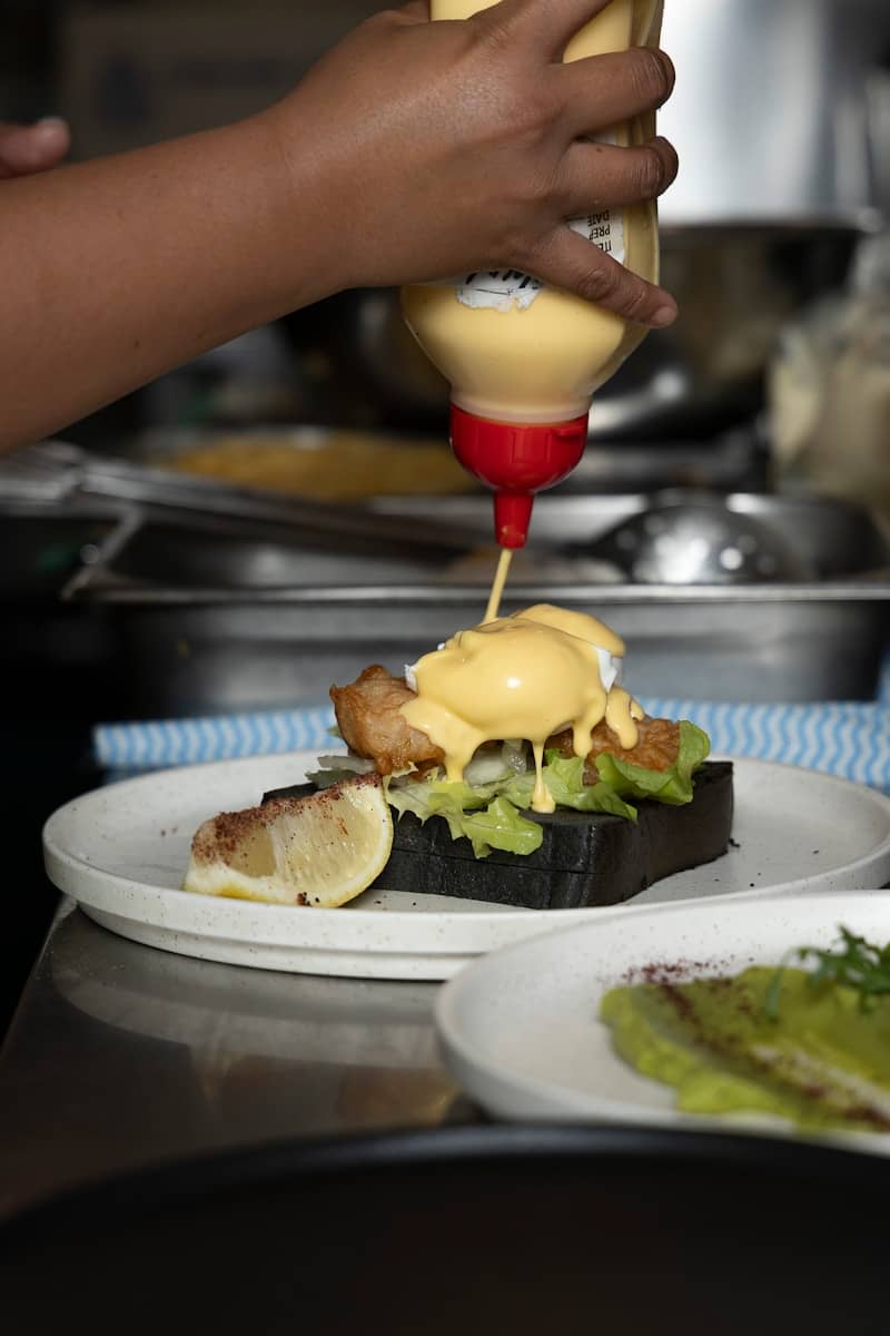 Sauce is being drizzled on an eggs benedict dish.